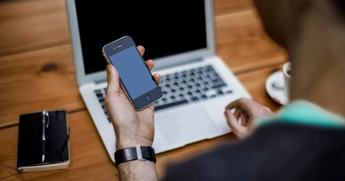 Une personne consulte son téléphone devant un ordinateur portable ouvert